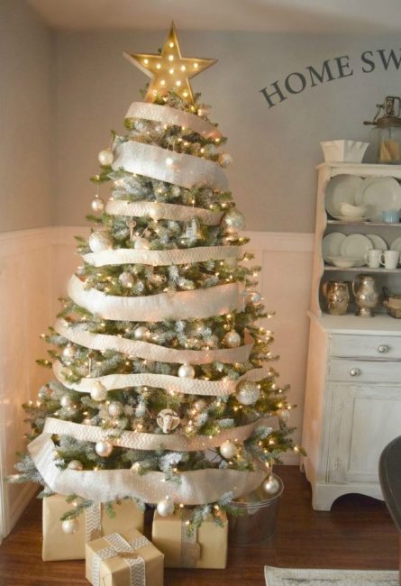 Ici, l'accent est mis sur le décor de rubans blancs et dorés, qui enveloppent généreusement le sapin de Noël en cercle.